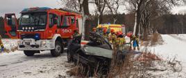 Samochód osobowy po uderzeniu w drzewo. W okolicy znajdują się pojazdy służb ratowniczych oraz strażacy. 