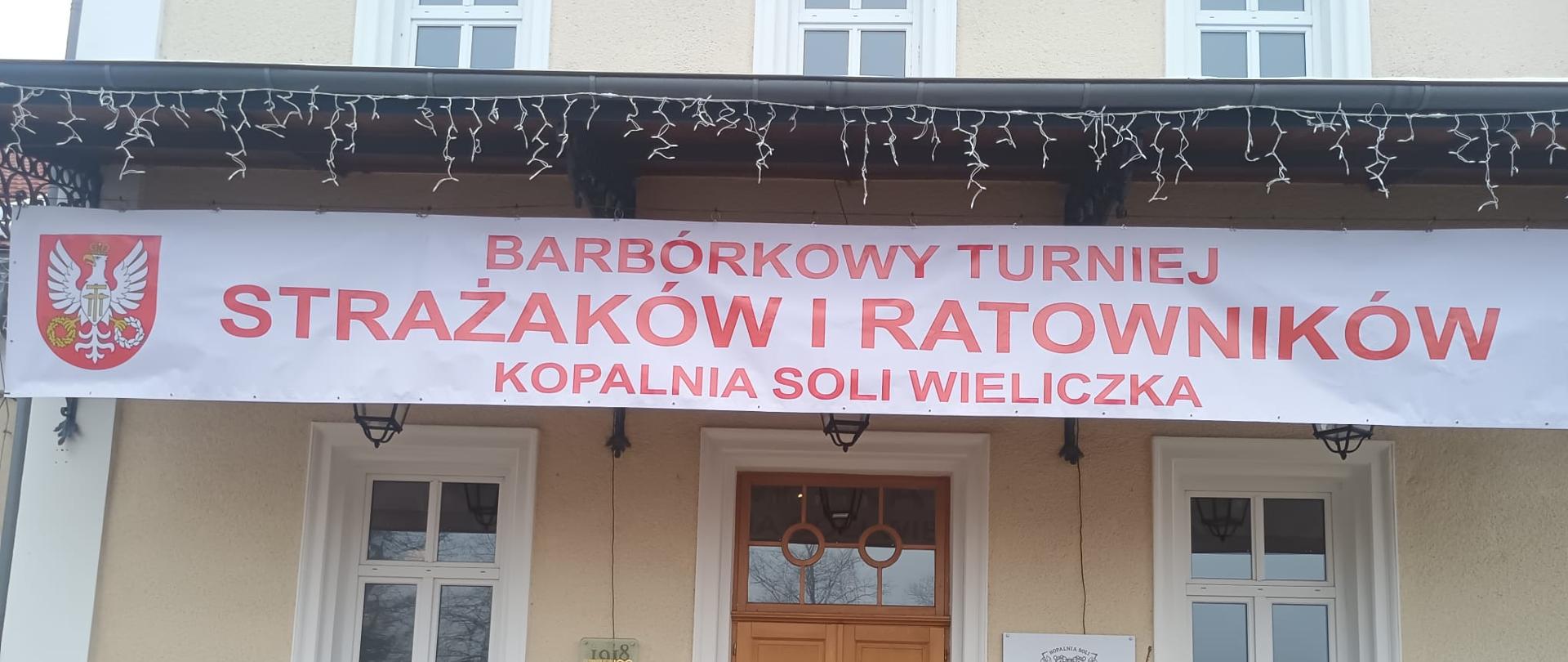 Zdjęcie przedstawia sześciu strażaków w umundurowaniu bojowym w nomeksach, czerwonych hełmach siedzących na schodach. W tle nad nimi wejście do Szybu Daniłowicza oraz napis nad nimi " Barbórkowy Turniej Strażaków i Ratowników Kopalnia Soli Wieliczka"
