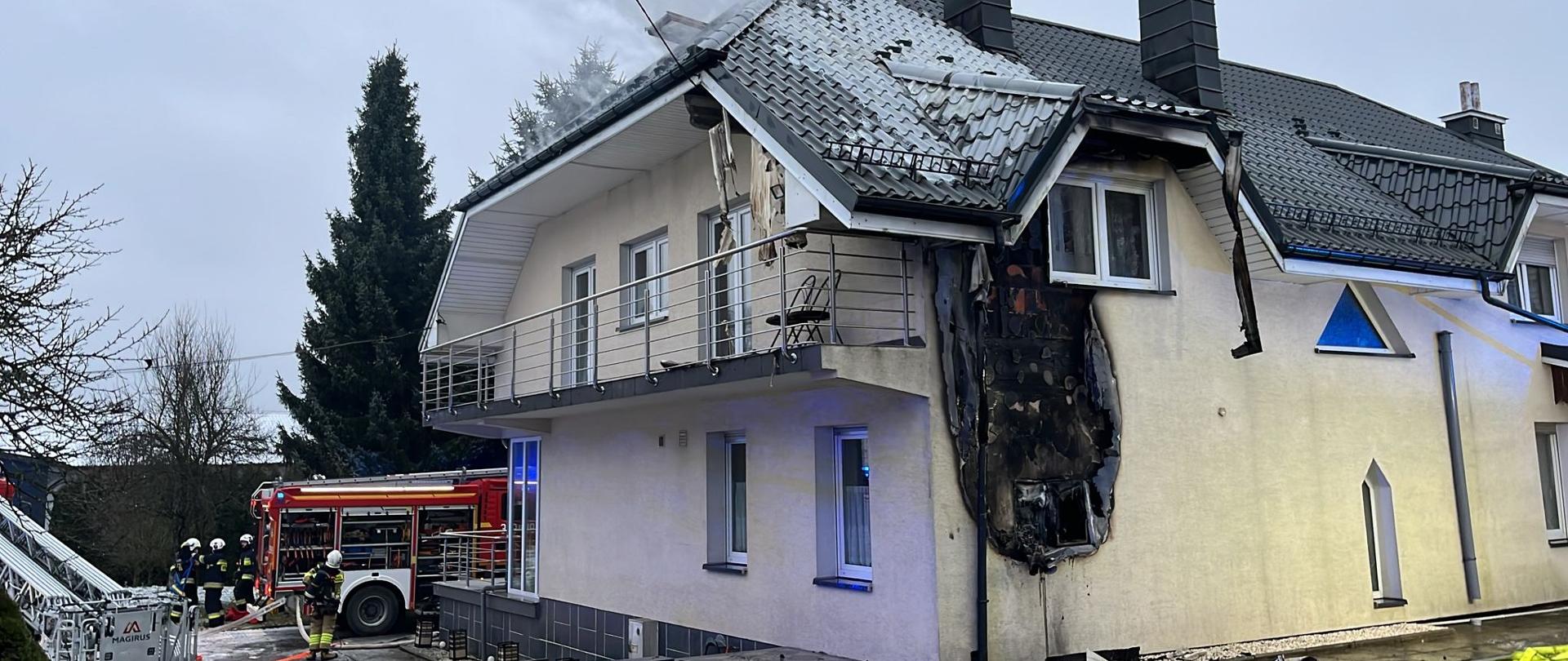 Zdjęcie przedstawia budynek w Masłowie gdzie doszło do pożaru elewacji i poddasza. W głębi pracują strażacy.