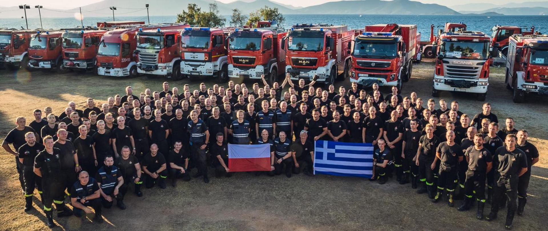Grupa polskich i greckich strażaków ze swoimi flagami państwowymi na tle samochodów pożarniczych