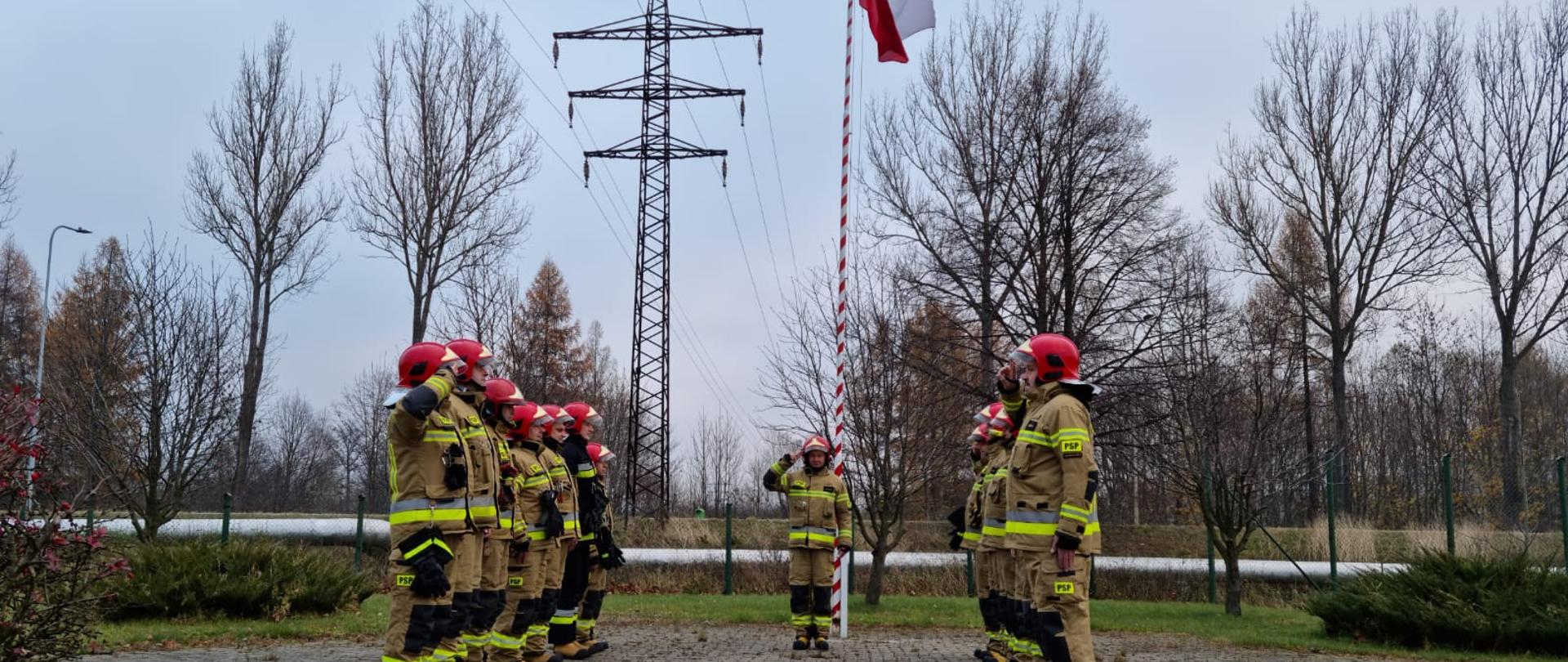 Uroczysta zmiana służby z okazji Obchodów Dnia Niepodległości przeprowadzana na zewnątrz budynku Komendy Powiatowej Państwowej Straży w Kamiennej Górze w tle masz flagowy, na którym powiewa flaga państwowa. Dowódca stojący po środku oraz dowódcy zmian salutują. Strażacy z poszczególnych zmian służbowych ustawieni w dwóch szeregach stoją na przeciw siebie. Wszyscy funkcjonariusze ubrani w ubrania specjalne koloru piaskowego.