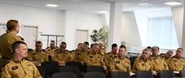 Kobieta strażak w umundurowaniu koloru musztardowego przemawia do kadry kierowniczej która siedzi na krzesłach. 