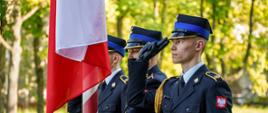 Podniesienie biało- czerwonej flagi na biało-czerwony maszt. Trzech strażaków w umundurowaniu wyjściowym. Środkowy kręcąc korbką wciąga flagę, dwaj pozostali (przyboczni) oddają honor przez salutowanie. W tle intensywna zieleń liści drzew. 