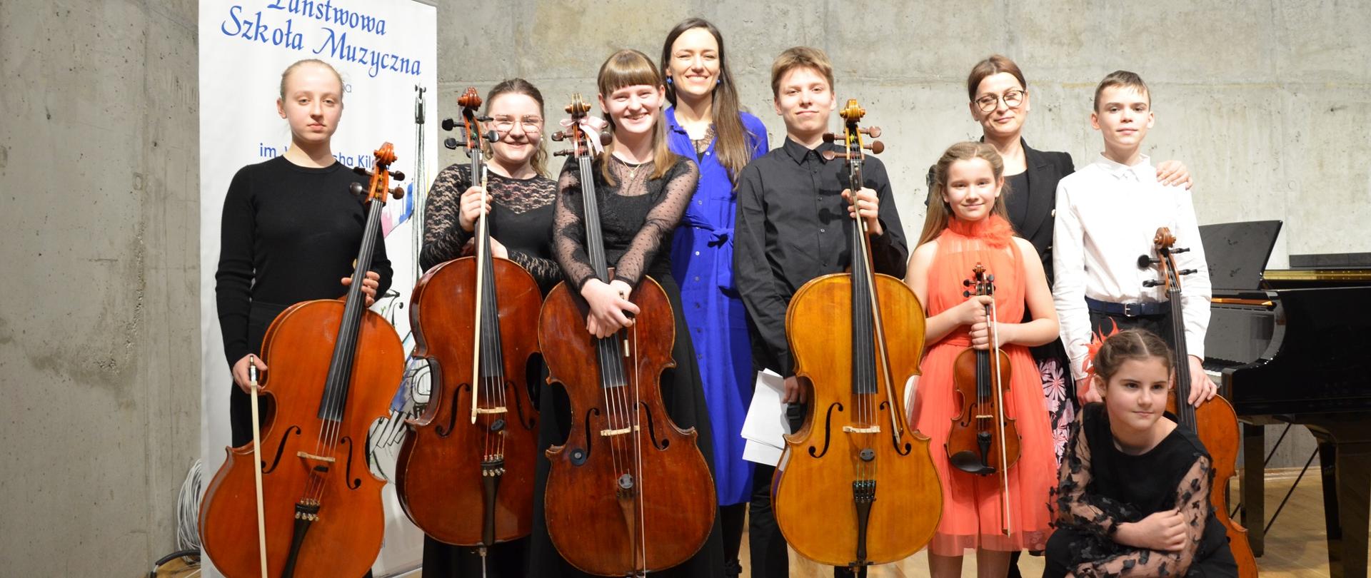 Grupa uczniów na scenie szkoły muzycznej, uczniowie trzymają wiolonczele i skrzypce, w tle baner promujący PSM w Wodzisławiu Śląskim