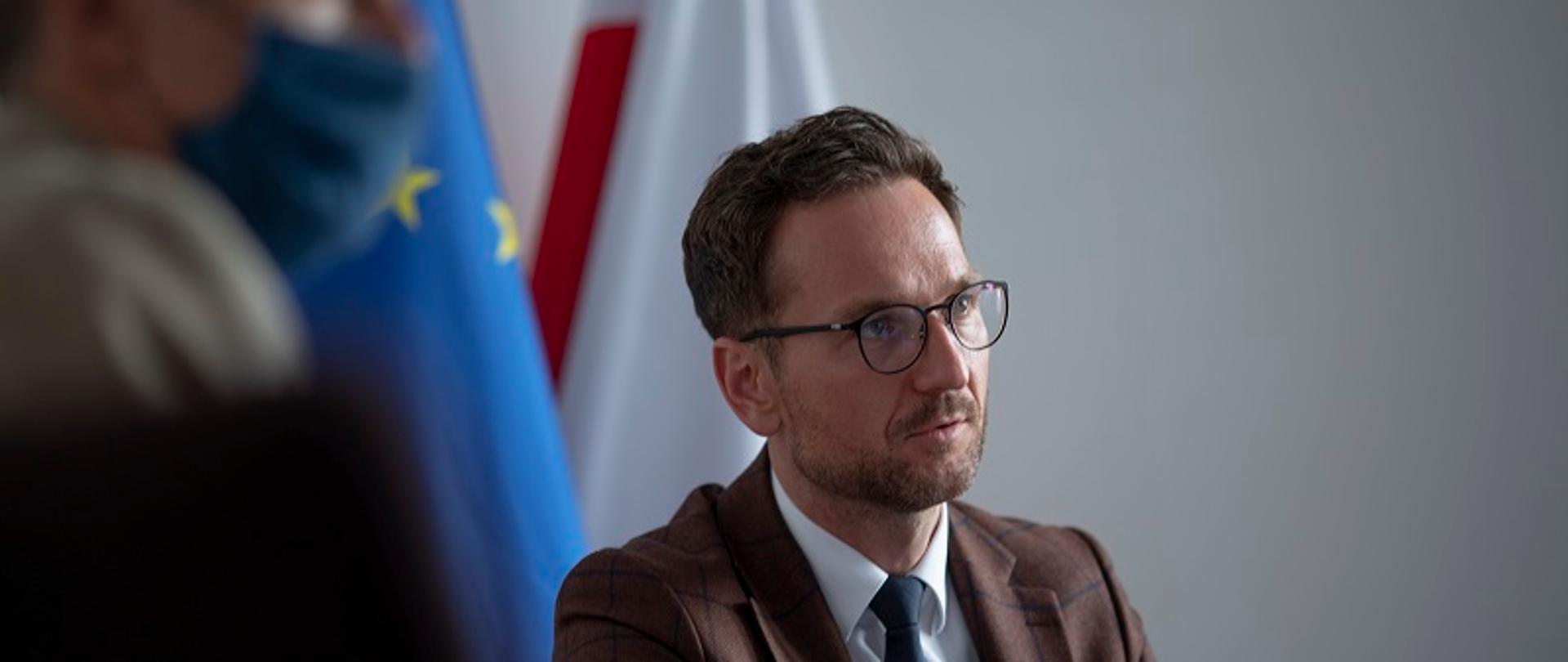 Minister Waldemar Buda siedzi przy stole konferencyjnym, za nim flagi Polski i UE.