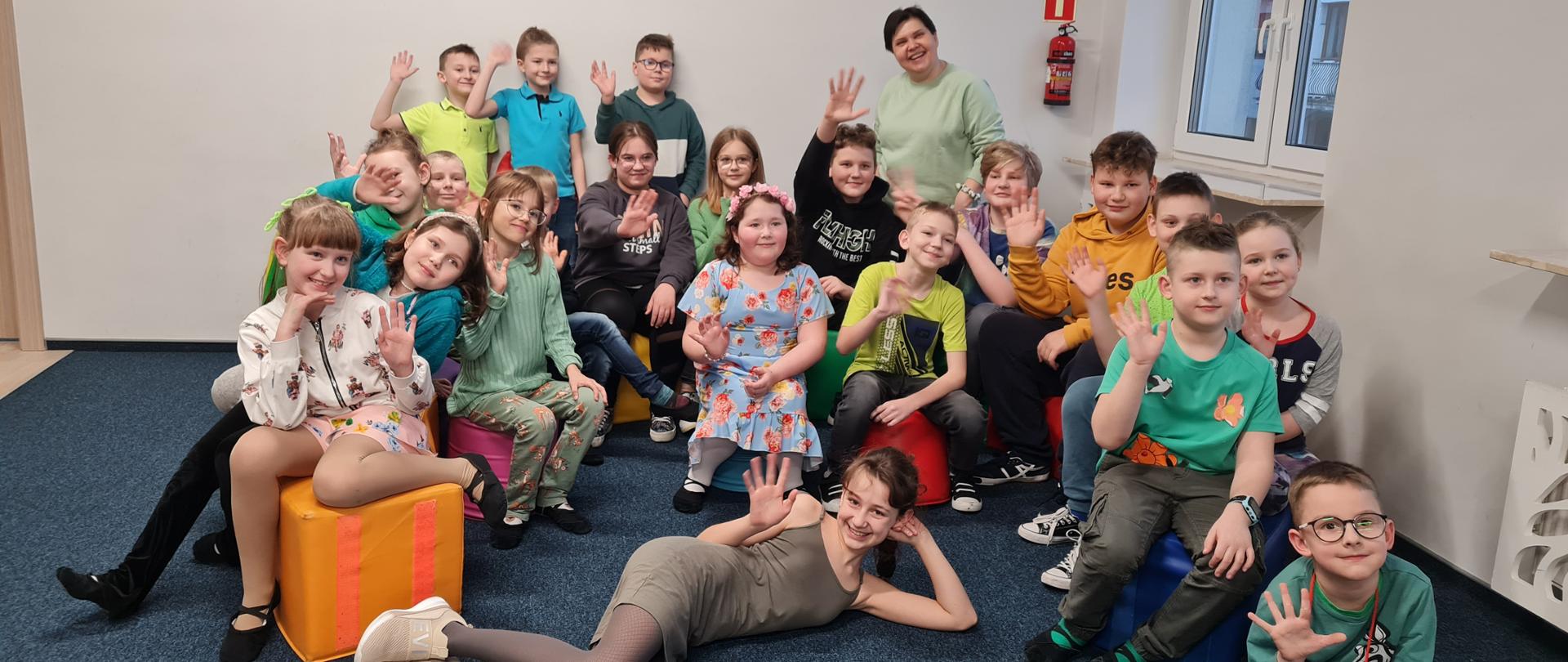 Zdjęcie przedstawia grupę uczniów z klasy I c.6 gr A i I c.4. Dzieci siedzą na pufach lub leżą przed nimi, są ubrane na kolorowo i machają. W tle okno a na podłodze granatowa wykładzina.