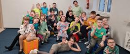 Zdjęcie przedstawia grupę uczniów z klasy I c.6 gr A i I c.4. Dzieci siedzą na pufach lub leżą przed nimi, są ubrane na kolorowo i machają. W tle okno a na podłodze granatowa wykładzina.