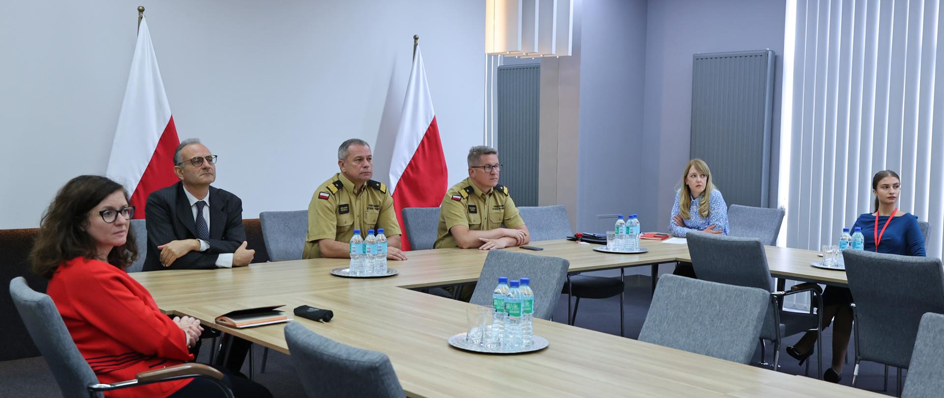 Trzej mężczyźni oraz trzy kobiety, siedzący przy stole konferencyjnym. Wszyscy patrzą w prawą stronę na coś poza kadrem z kadrem. 