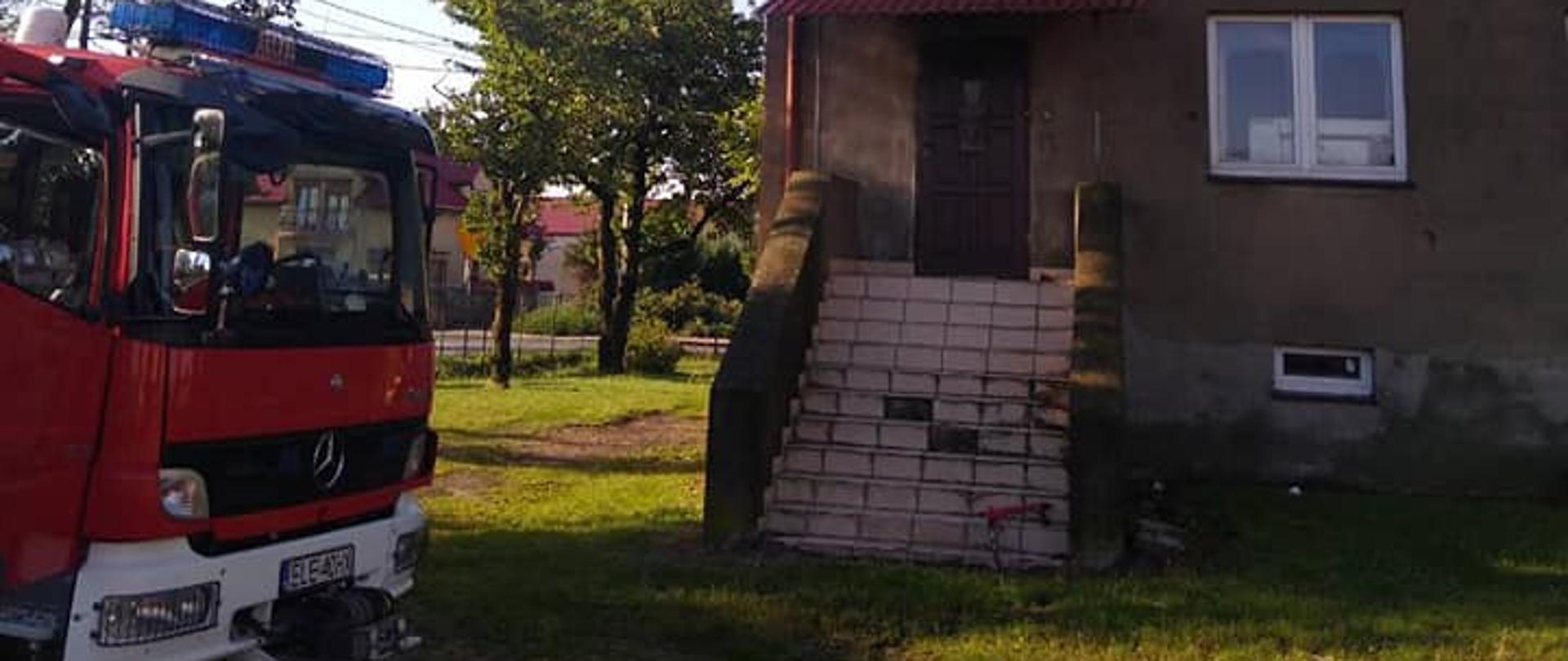 Na zdjęciu widać piętrowy budynek mieszkalny wielorodzinny, w którym doszło do wybuchu gazu. W budynku część okien jest pootwierana. W jednym oknie są wybite szyby. Przed budynkiem po lewej stronie stoi czerwony samochód strażacki. W tle widać inne budynki oraz drzewa. Jest widno.