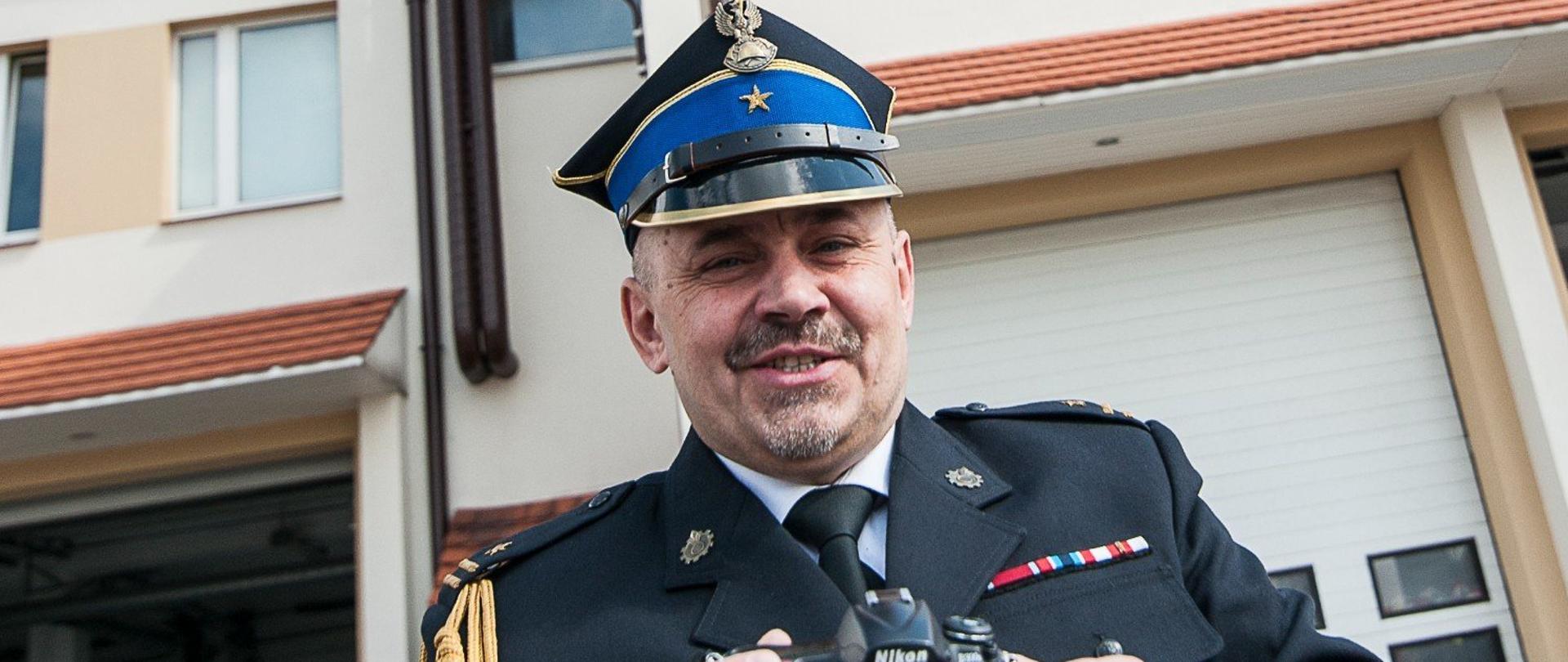 uśmiechnięty strażak w rogatywce, mundurze ze sznurem trzyma w ręku aparat, w tle brama garażowa 