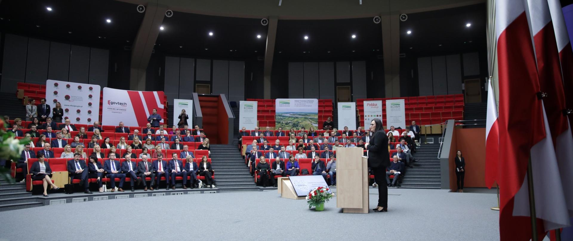 Na zdjęciu aula, wokół której siedzą osoby. Na środku mównica, z której przemawia minister Małgorzata Jarosińska-Jedynak.