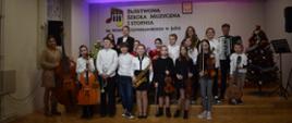 Grupa uśmiechniętych uczniów stojąca na scenie auli PSM trzymająca swoje instrumenty i pozująca do zdjęć. W tle logo szkoły, Godło, ogólna kolorystyka – barwy ciepłe.