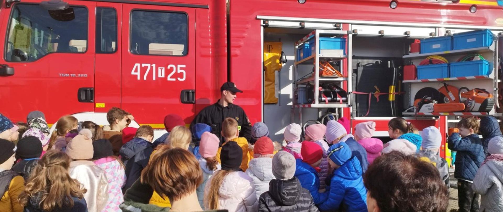 Zdjęcie zrobione w dzień. Strażaka stojący przed autem pożarniczym przedstawiający sprzęt ratowniczy zgromadzonym uczniom.