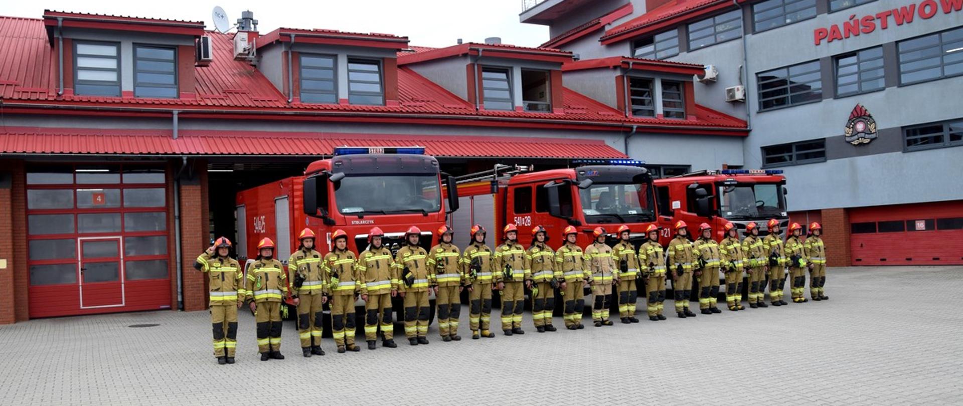 Zdjęcie przedstawia strażaków w umundurowania specjalnym, którzy stoją w szeregu na zbiórce. W tle samochody strażackie i budynek Komendy.