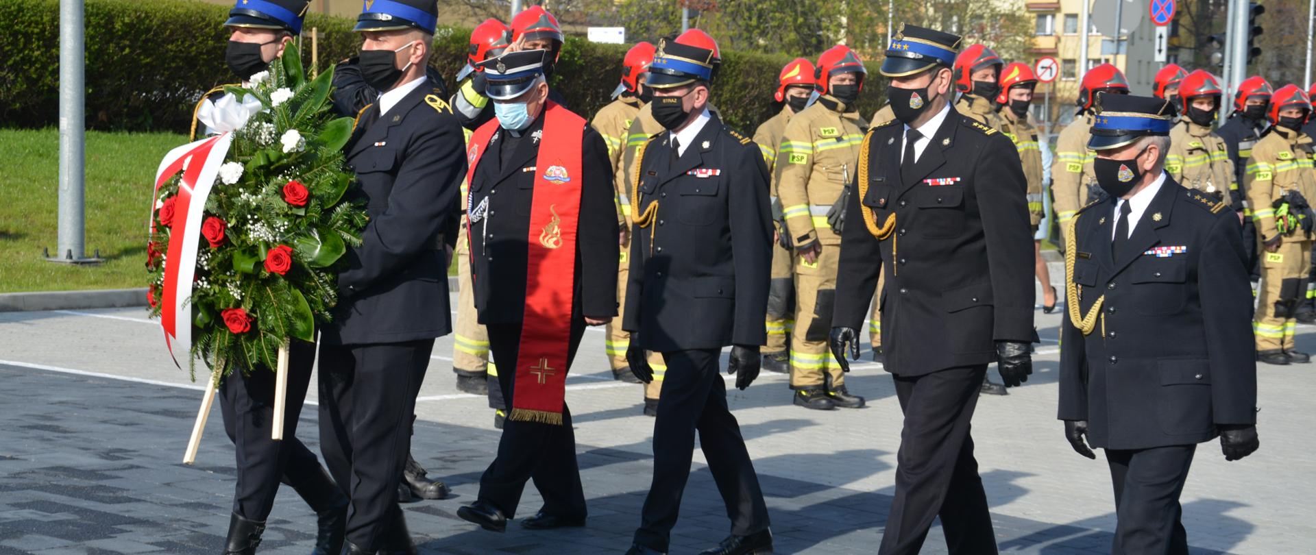 Zdjęcie przedstawia dwóch strażaków niosących wiązankę kwiatów. Za nimi w szeregu idzie kapelan strażaków, komendant miejski Państwowej Straży Pożarnej oraz świętokrzyski komendant wojewódzki Państwowej Straży Pożarnej wraz ze swoim zastępcą. W tle widać bloki mieszkalne oraz strażaków stojących w dwuszeregu ubranych w ubrania bojowe.