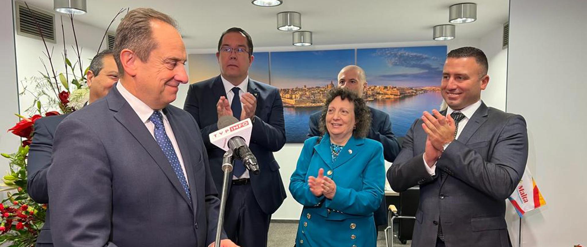 Na zdjęciu znajdują się elegancko ubrani mężczyźni i kobieta. Jeden z mężczyzn jest w trakcie wystąpienia publicznego. 