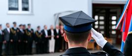 Zdjęcie przedstawia strażaka Państwowej Straży Pożarnej w galowym mundurze, widzianego od tyłu podczas wykonywania salutowania. W tle znajduje się budynek jednostki PSP oraz zebrani goście i funkcjonariusze, co wskazuje na uroczysty charakter wydarzenia. Mężczyzna nosi czapkę rogatywkę i białe rękawiczki, co sugeruje udział w oficjalnej ceremonii – prawdopodobnie promocji oficerskiej. Kadrowanie i wyrazisty gest nadają zdjęciu podniosły, patriotyczny charakter. Scena uchwycona została w momencie pełnym powagi i szacunku.