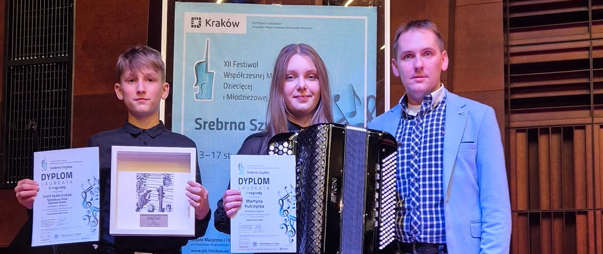 Zdjęcie przedstawia trzy osoby stojące obok tablicy informacyjnej konkursu "Srebrna Szybka". Po prawej stoi dorosły mężczyzna w błękitnej marynarce i jeansach. Obok niego młoda kobieta trzymająca czarny akordeon oraz dyplom. Z przodu klęczy chłopiec, który trzyma w rękach dyplom oraz statuetkę w ramce. W tle widoczna jest drewniana ściana sali koncertowej.