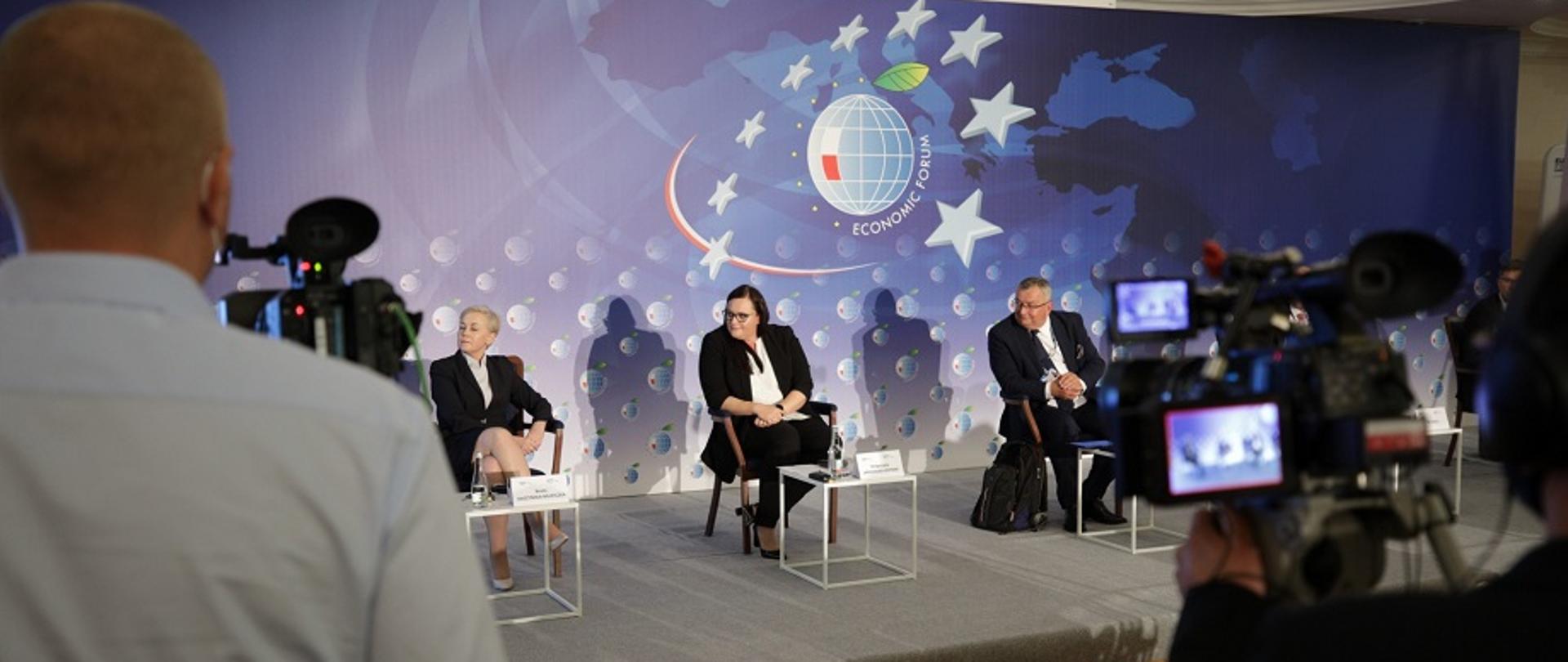 Na zdjęciu scena i troje panelistów siedzących, dwie kobiety i mężczyzna. Jedną z kobiet jest minister Małgorzata Jarosińska-Jedynak. Za plecami ścianka z logo i napis Economic Forum. Na pierwszym planie dwóch operatorów kamer.
