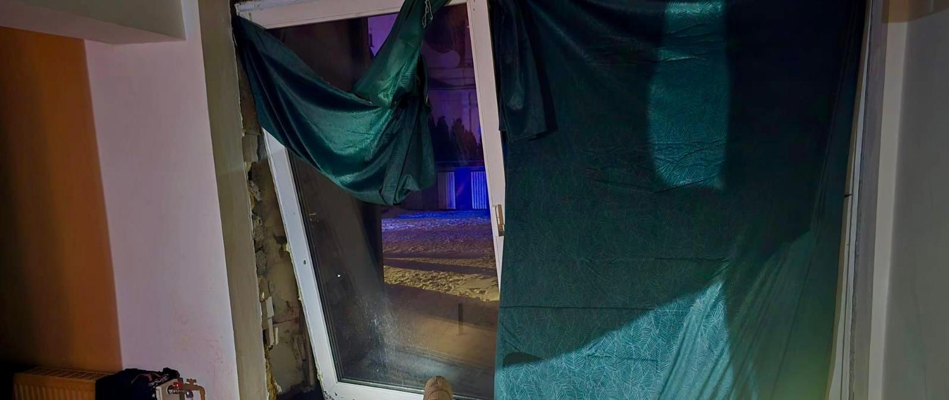 Wnętrze pokoju z otwartym oknem, uszkodzonym oknem zasłoniętym zieloną tkanioną; na stole talerz z jedzeniem i kubek, obok grzejnik i krzesło, za oknem widać ośnieżoną ulicę nocą. 