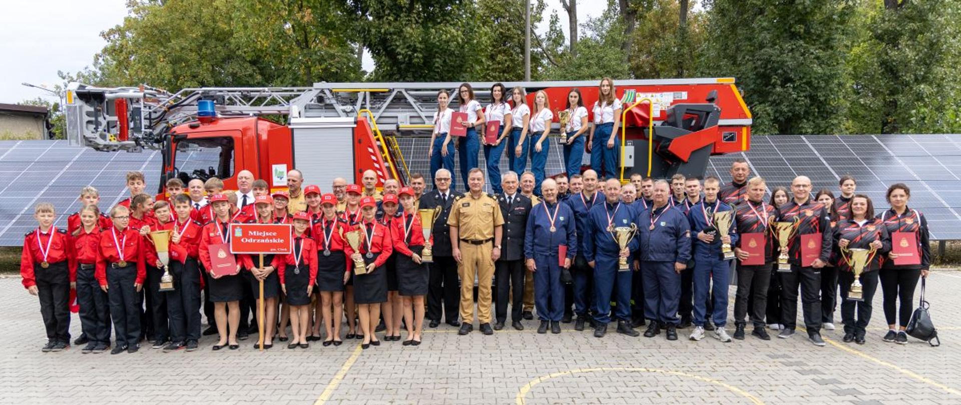 Uroczysta zbiórka jednostek OSP na placu Komendy Wojewódzkiej PSP w Opolu – druhny i druhowie w grupie, na tle samochodu pożarniczego z drabiną mechaniczną. Wspólne zdjęcie grupowe uczestników zbiórki.