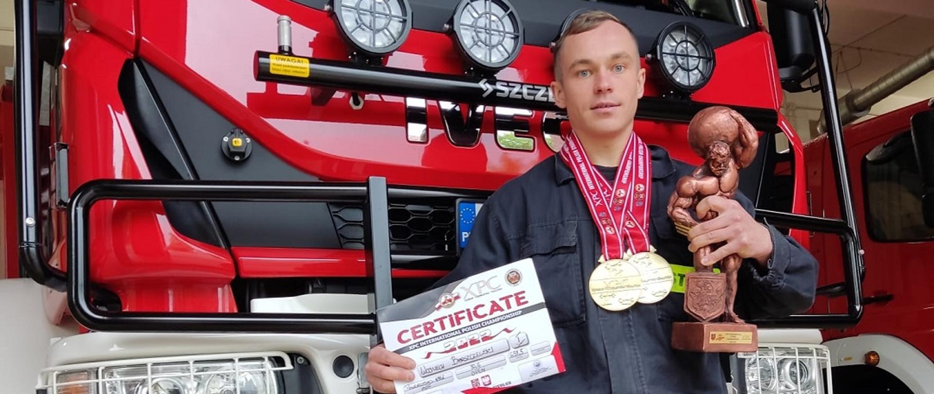 Zdjęcie obrazuje strażaka z medalami w rekach trzyma statuetkę dyplom w tle pojazd pożarniczy