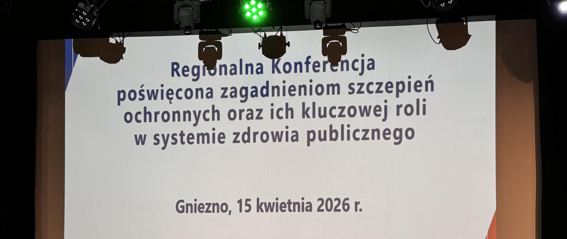 Duży ekran projekcyjny na scenie z wyświetloną planszą konferencyjną. Na górze kadru widoczne są reflektory sceniczne, w tym jedno zielone światło świecące na środku; na dole widać ciemną podłogę sceny. Tekst na ekranie (verbatim): Regionalna Konferencja poświęcona zagadnieniom szczepień ochronnych oraz ich kluczowej roli w systemie zdrowia publicznego Gniezno, 15 kwietnia 2026 r. Na dole znajdują się logotypy; po lewej czerwony kwadrat z białymi znakami „e*s +te de)”, pośrodku dwa okrągłe znaki i napis „CHRONIMY ZDROWIE Z MYŚLĄ O PRZYSZŁOŚCI”, po prawej okrągła pieczęć z drobnym tekstem (nieczytelnym w tym ujęciu).