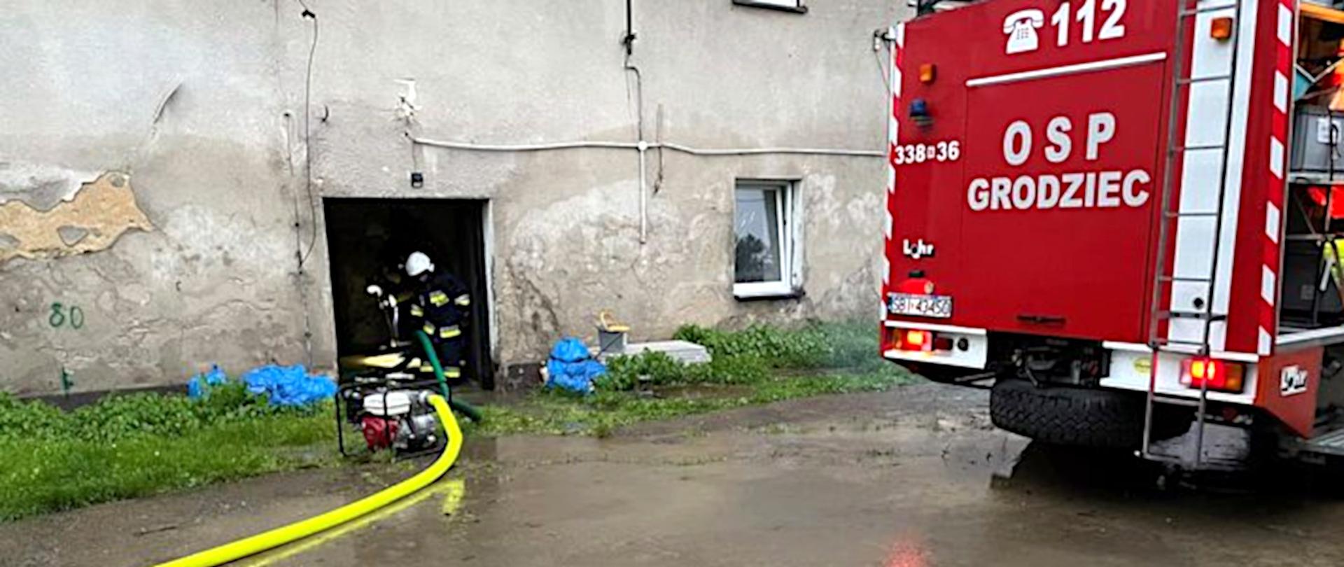 Na zdjęciu widoczna jest zalana wodą ulica przed budynkiem. Po prawej stronie stoi czerwony samochód specjalny OSP. Po lewej stronie w drzwiach budynku stoi przy motopompie dwóch strażaków w ubraniach specjalnych. Strażacy wypompowują wodę z piwnicy budynku.