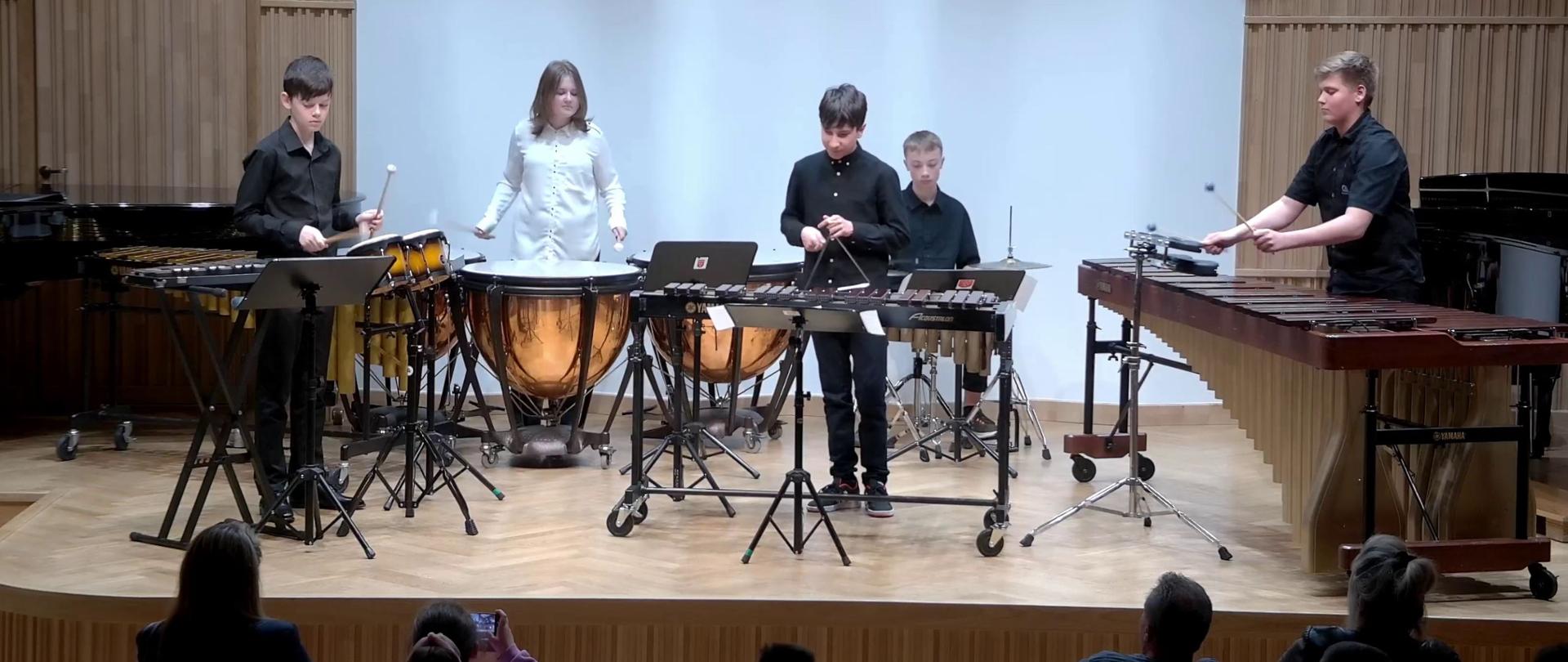 Zdjęcie. Na estradzie sali koncertowej, przed publicznością, występuje zespół perkusyjny. Od lewej strony uczniowie grają na: bongosach, kotłach, trójkącie, zestawie perkusyjnym i marimbie.
