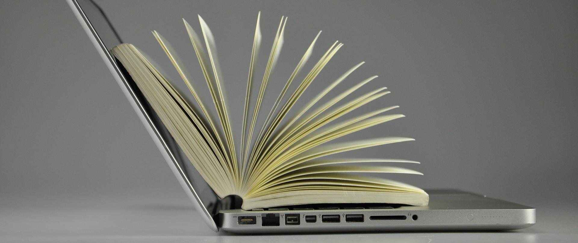 Na zdjęciu znajduje się laptop, na którym leży otwarta książka.