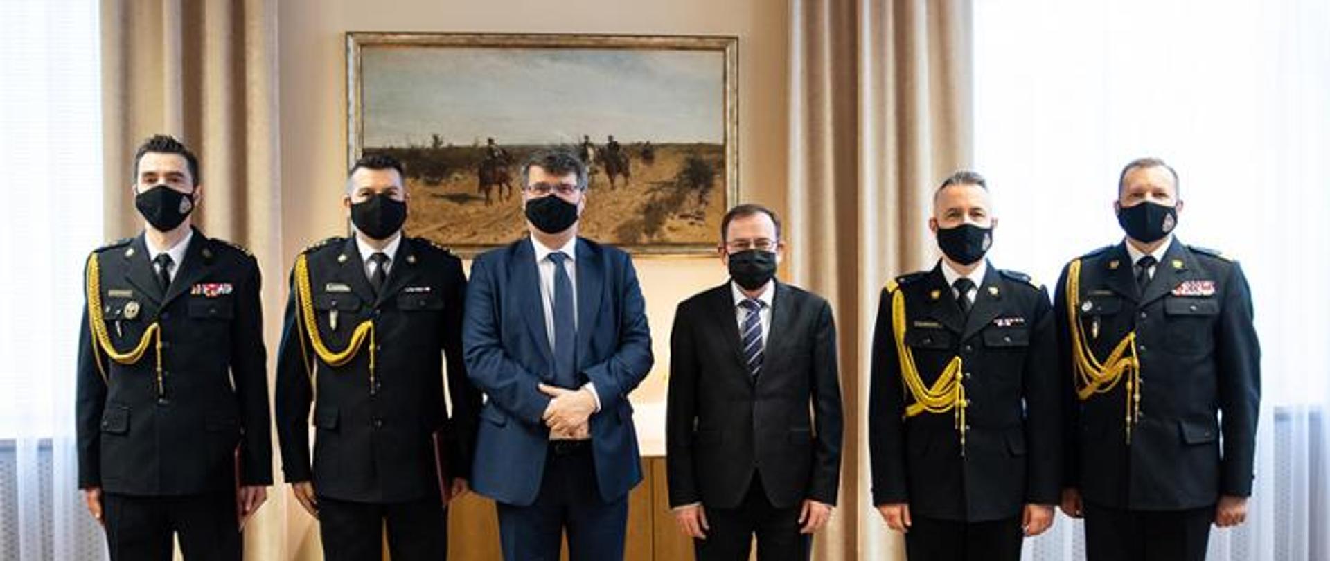 Od lewej stoją: st. bryg. Mariusz Feltynowski,st. bryg. Jarosław Chodorowski, minister Macej Wąsik, minister Mariusz Kamiński, nadbryg. Andrzej Bartkowiak oraz nadbryg Adam Konieczny. W tle ściana udekorowana obrazem.
