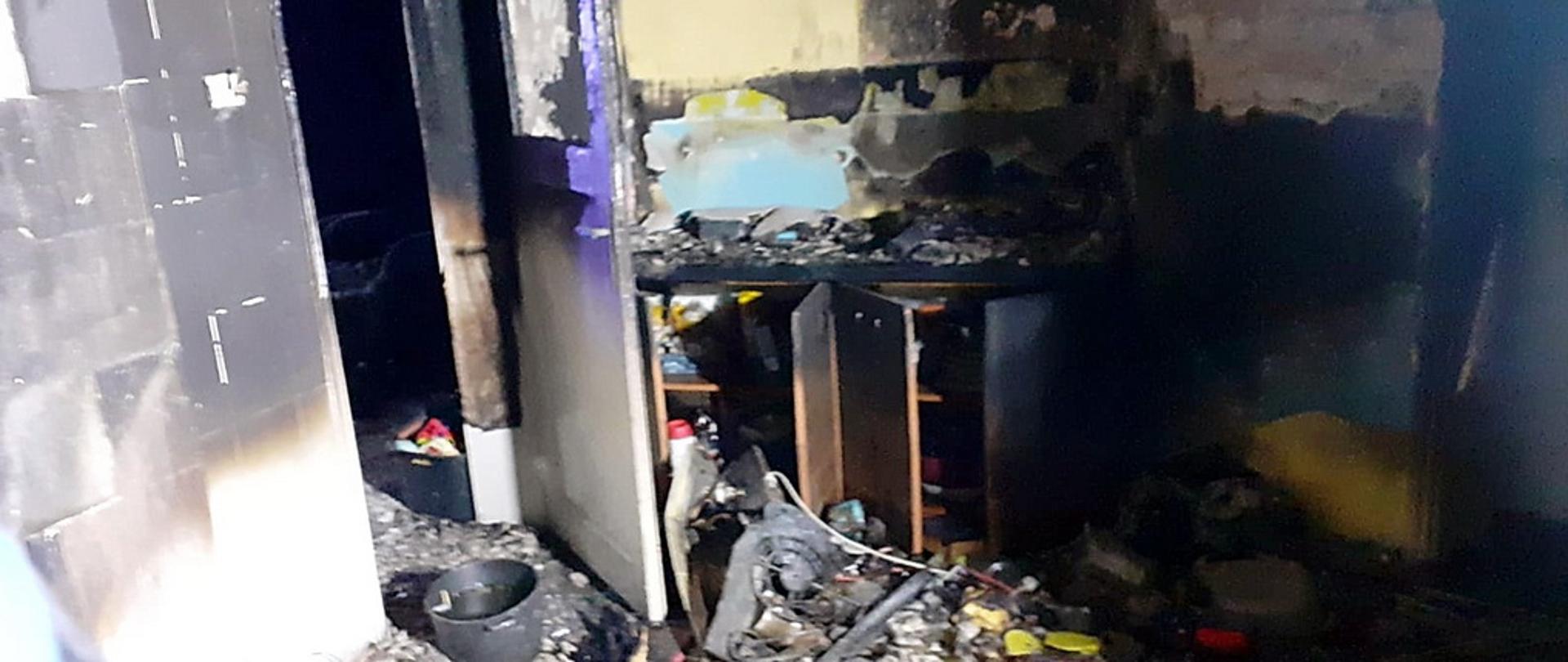 Wnętrze spalonej kuchni w budynku mieszkalnym, ściany opalone aż do cegły, na podłodze widoczne spalone szczątki wyposażenia i mebli, w tle widoczne nadaplone drzwi i szafka kuchenna