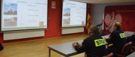 Konferencja upamiętniająca pożar lasu w Kuźni Raciborskiej. Świetlica Komendy Powiatowej PSP w Rawiczu. Przy stołach siedzą strażacy w ubraniach koszarowych. Są zwróceni w stronę ekranów, na których oglądają przebieg konferencji i wyświetlaną prezentację multimedialną. Pomiędzy ekranami zawieszone jest godło państwowe. Z prawej strony widoczny jest stojak z flagami oraz ścianka promocyjna.