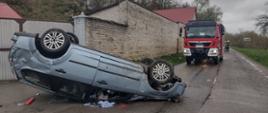 Pojazd przewrócony na dach, wybite szyby, resztki powypadkowe porozsypywane wokół. Za pojazdem samochód gaśniczy i strażacy zabezpieczający miejsce zdarzenia.