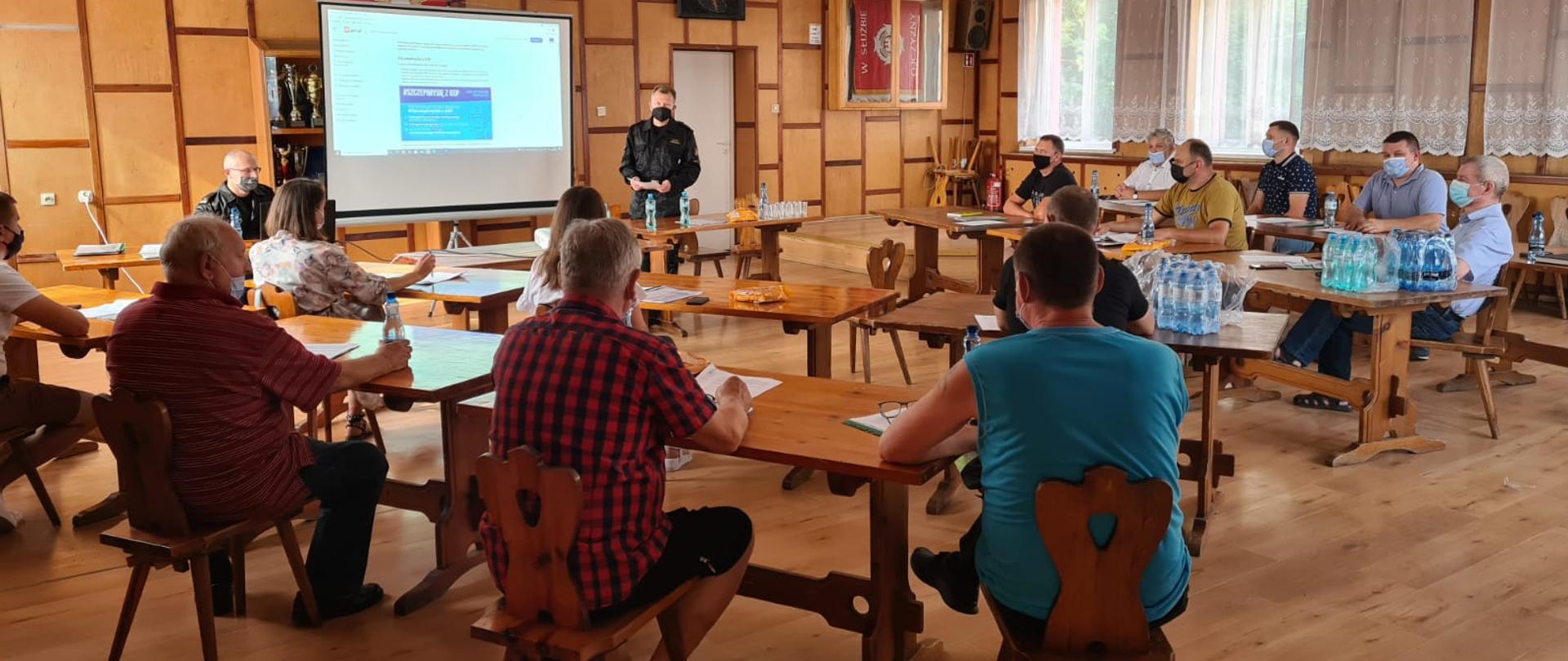 zdjęcie zrobione na świetlicy OSP w Kamieniu Krajeńskim. Przodem do aparatu stoi Komendant Powiatowy PSP st. bryg. Maciej Suchomski, obok przy komputerze siedzi Zastępca Komendanta mł. bryg. Dariusz Chylewski. Pomiędzy nimi stojak na którym wyświetlony jest slajd programu SczepimySię z OSP. Tyłem przy rozstawionych stolikach po lewej stronie patrzącego siedzą druhowie (przedstawiciele OSP) oraz koordynatorzy gminni ds. szczepień przeciwko Covid-19. Przodem po prawej stronie patrzącego siedzą druhowie (przedstawiciele OSP) oraz koordynatorzy gminni ds. szczepień przeciwko Covid-19