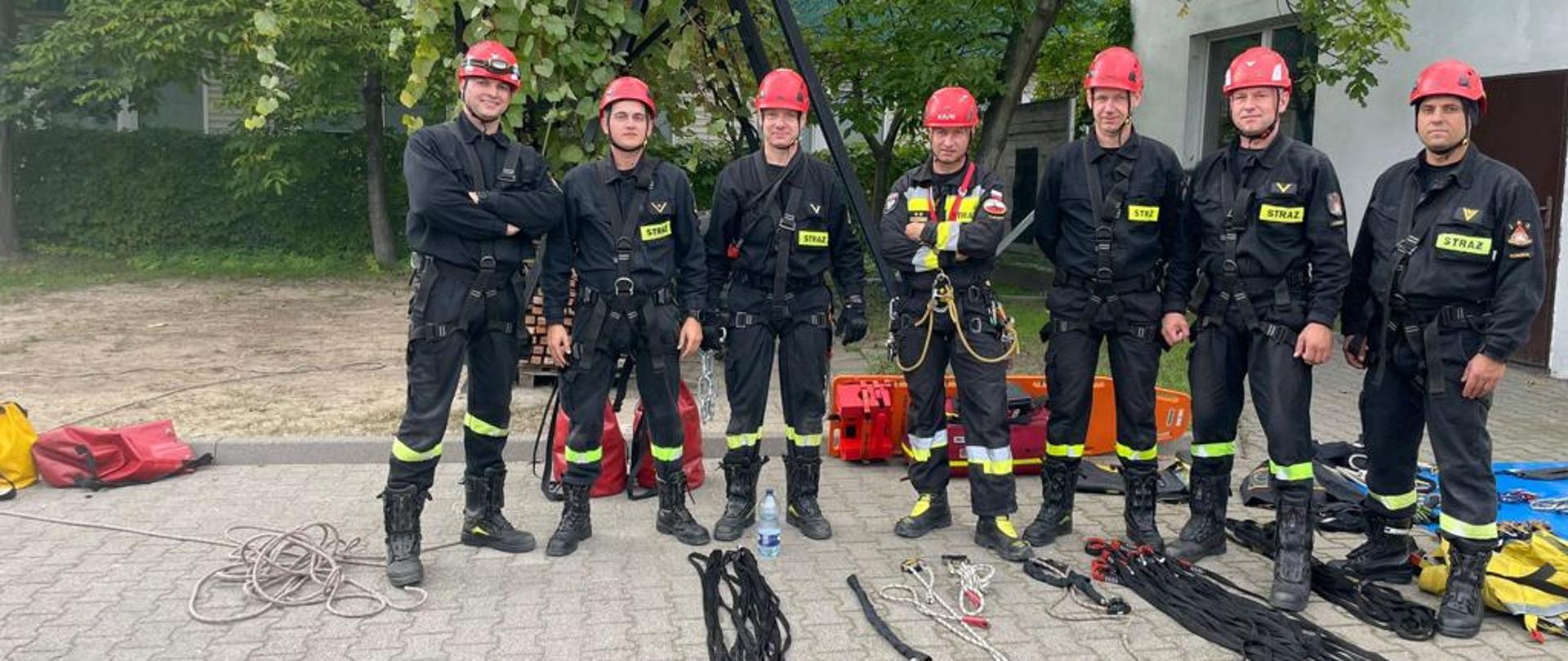 Grupa strażaków stoi w jednym rzędzie, przed nimi leży sprzęt przygotowany do ćwiczeń z ratownictwa wysokościowego