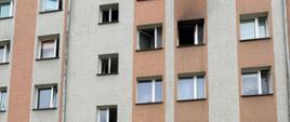 Blok wielorodzinny z widocznymi uszkodzeniami oraz wydzielonym miejscem za pomocą stożków ostrzegawczych i taśmy do ewakuacji nadpalonych elementów wyposażenia mieszkania.