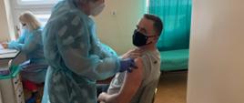 Zdjęcie przedstawia mężczyznę siedzącego na krześle z odsłoniętym ramieniem, który jest szczepiony, przez pielęgniarkę. 