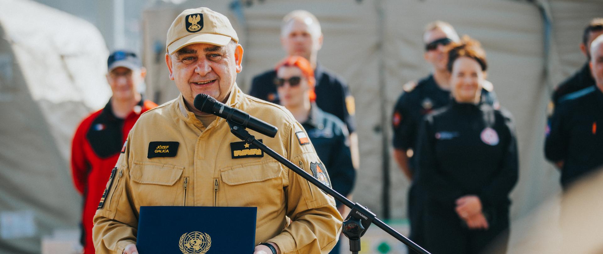 Józef Galica w trakcie przemówienia przy mikrofonie. W tle stoją ludzie oraz rozstawione są namioty. 