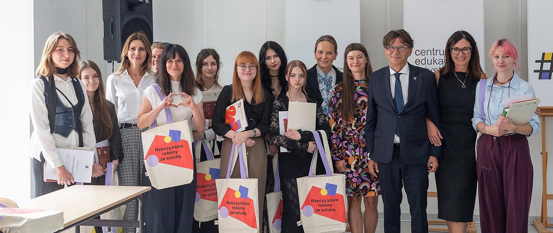 Pionowe zdjęcie grupowe zrobione w nowych pracowniach. Większość obecnych na zdjęciu osób to młode kobiety ubrane w eleganckie oraz półformalne stroje, trzymające płócienne torby i dyplomy lub dokumenty. Po prawej stronie stoi mężczyzna w ciemnym garniturze i okularach z jedną z kobiet opierającą o niego dłoń. Tło tworzą białe ścianki z zawieszonymi dużymi literami oraz tablica edukacyjna.