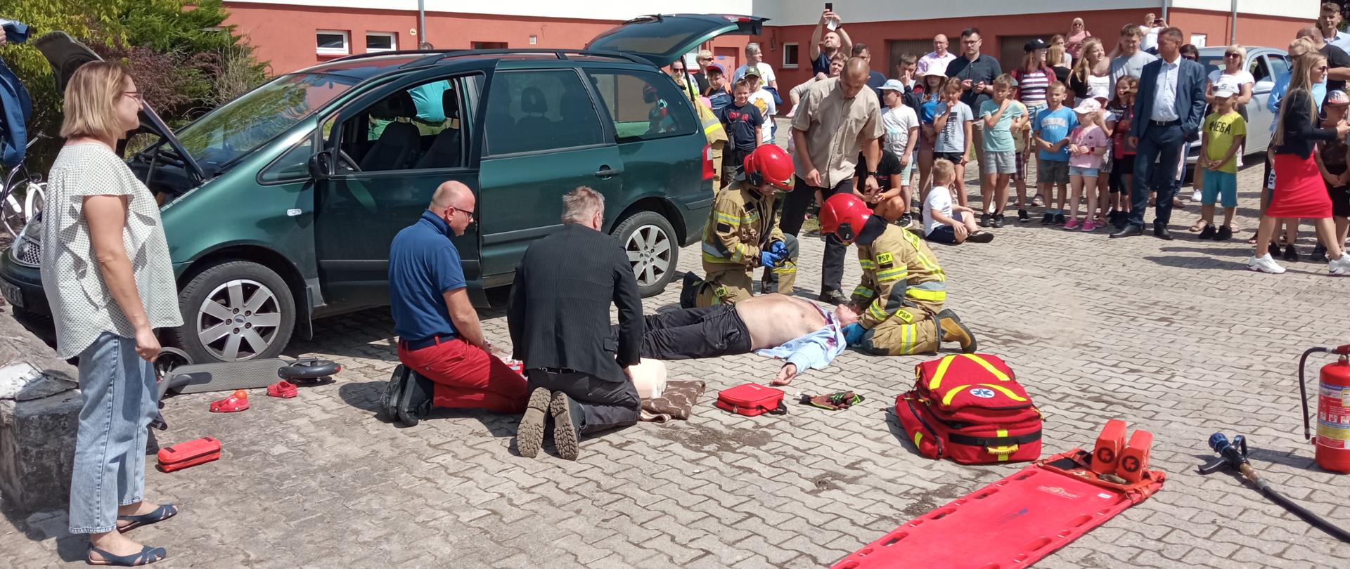 Zdjęcie przedstawia ćwiczenia dot. udzielania pierwszej pomocy medycznej w związku ze zdarzeniami w transporcie komunikacyjnym. Obraz przedstawia dwóch strażaków Komendy Powiatowej Państwowej Straży Pożarnej w Gołdapi zajmujących się jedna z osób poszkodowanych oraz jednego strażaka, który przeszukuje pojazd osobowy pod kątem obecności innych podróżujących. Na fotografii widać również torbę medyczną, deskę ortopedyczną oraz zabezpieczenie gaśnicze pojazdu w postaci gaśnicy i nawodnionej linii gaśniczej. Na obrazie widać również dwie osoby (uczestnicy szkolenia), które udzielają pomocy medycznej kolejnej osobie poszkodowanej. Z lewej strony znajduje się osoba zgłaszająca zdarzenie. W tle natomiast znajdują się obserwatorzy m.in. z Ochotniczych Hufców Pracy