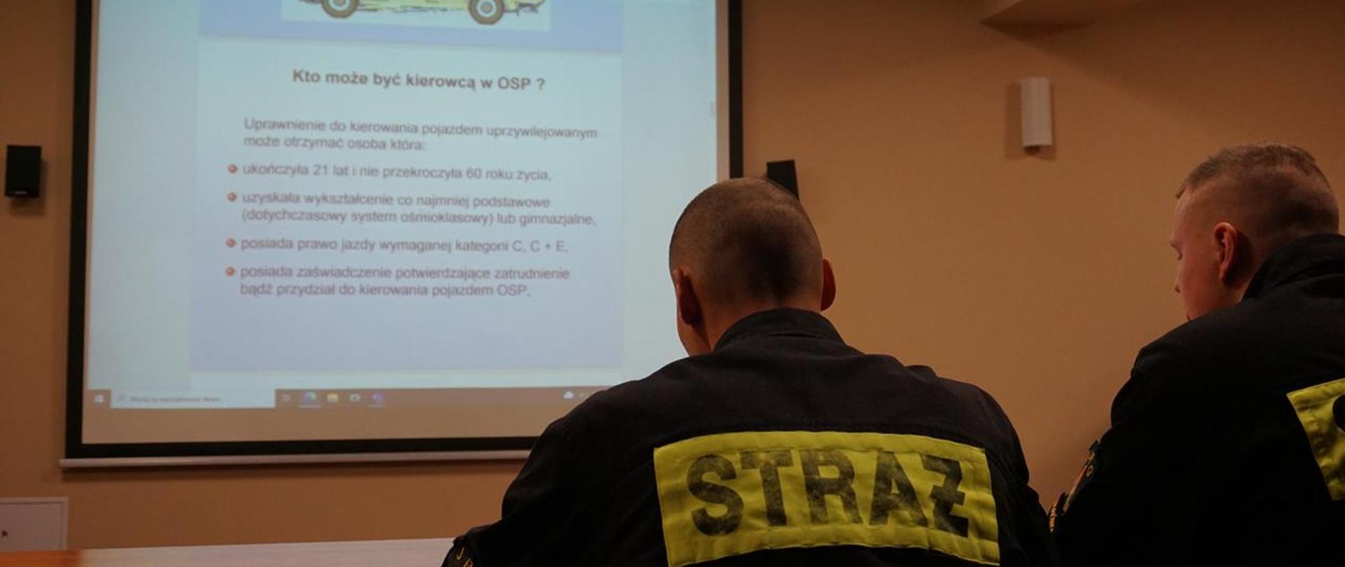 Dwóch strażaków w czarnych mundurach, odwróconych tyłem patrzą na prezentację multimedialną na ekranie.