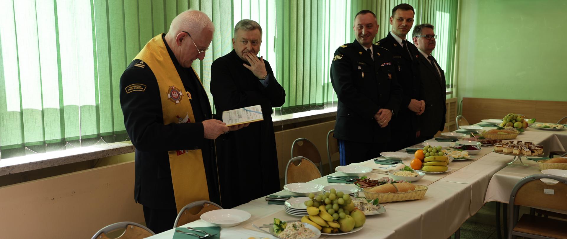 Zdjęcie przedstawia moment spotkania wielkanocnego w Szkole Aspirantów PSP w Krakowie, którym jest poświęcenie przez dwóch stojących księży darów. Koło księży stoi dwóch funkcjonariuszy w galowych mundurach i jedna osoba w garniturze Zdjęcie wykonane w pomieszczeniu z jasnozielonymi ścianami i dużymi oknami zasłoniętymi zielonymi pionowymi żaluzjami. Na stole przed nimi znajdują się półmiski z potrawami wielkanocnymi: jajka, owoce, wędliny, pieczywo, sałatki i ciasta. Obok nich – talerze, szklanki oraz zielone serwetki. W tle kilku mężczyzn w mundurach i garniturach siedzi i przysłuchuje się wystąpieniu. Atmosfera na zdjęciu jest spokojna i uroczysta – uczestnicy są skupieni, a stół symbolicznie nawiązuje do wspólnego świętowania Wielkiej Nocy.