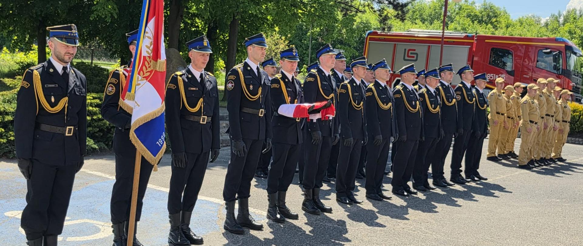 Uroczysty apel z okazji Dnia Strażaka przed siedzibą KP PSP w Kłobucku. W szeregu stoją strażacy Państwowej Straży Pożarnej w galowych mundurach, obok pocztu sztandarowego z biało-czerwonym sztandarem. Jeden ze strażaków trzyma złożoną flagę państwową. W tle widoczny jest czerwony samochód pożarniczy oraz grupa młodzieży w umundurowaniu. Uroczystość odbywa się w słoneczny dzień na placu przed jednostką, w otoczeniu zielonych drzew