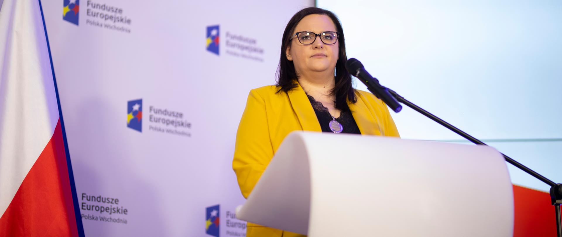 Na zdjęciu wiceminister Małgorzata Jarosińska-Jedynak stoi przy mównicy, obok flaga RP, z tyłu jasne tło