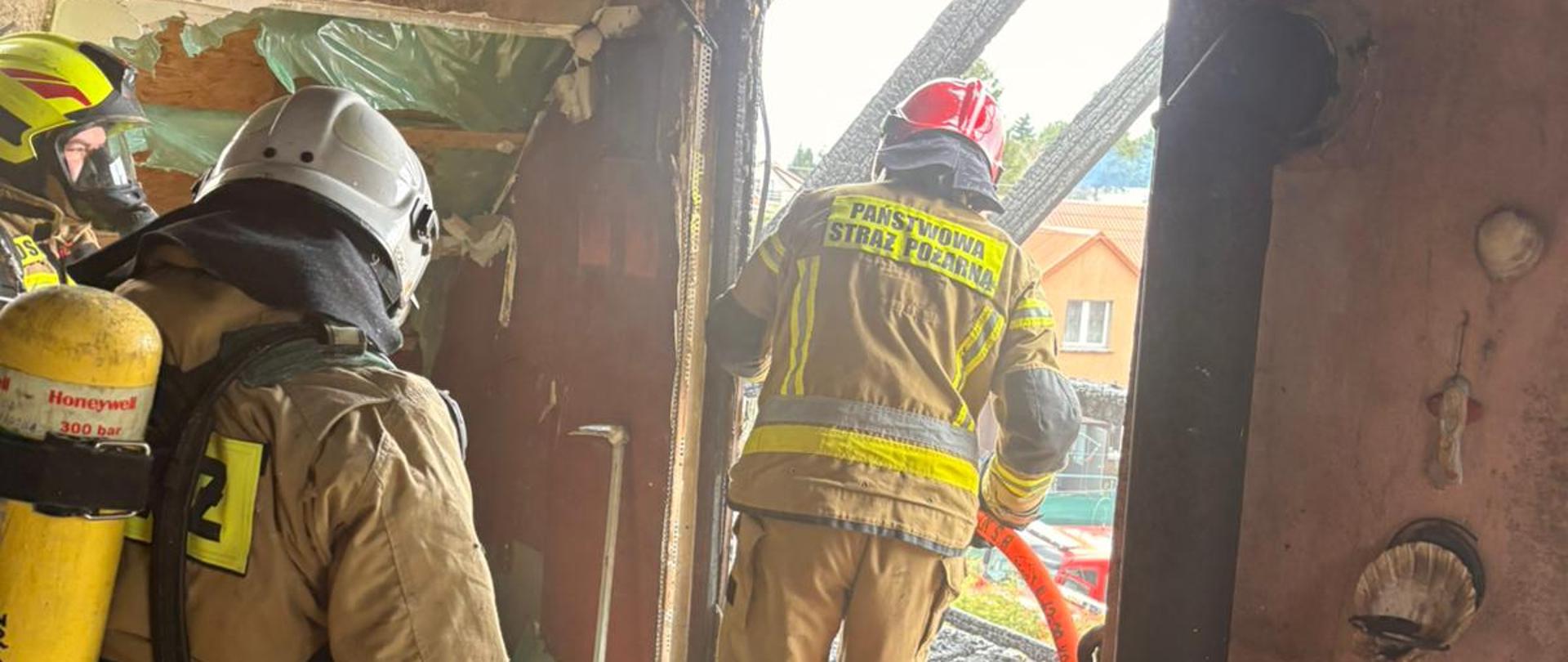 Widok z góry na akcję strażacką przy pożarze domu jednorodzinnego w zabudowie osiedlowej. Na ulicy stoją trzy czerwone wozy strażackie i samochód z drabiną, a strażacy prowadzą działania gaśnicze, rozwijając linie wężowe w kierunku nadpalonego budynku. Wokół znajdują się ogrody, ogrodzenia oraz sąsiednie domy jednorodzinne.