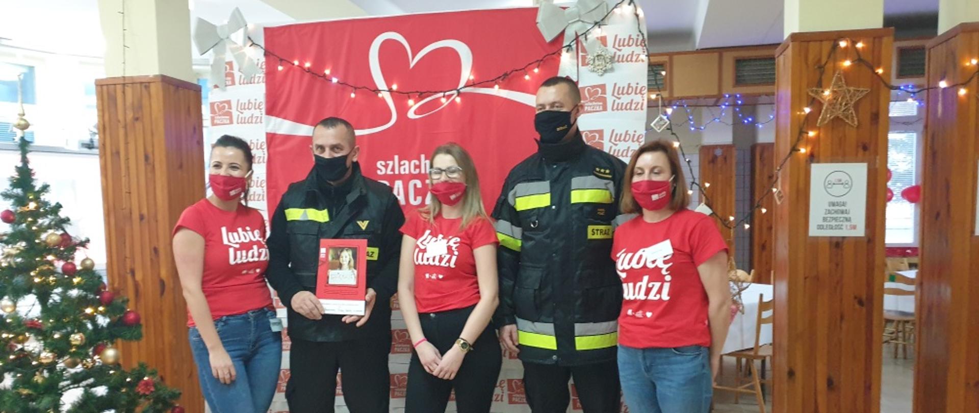 Zdjęcie przedstawia dwóch strażaków w umundurowaniu służbowym trzymających dyplom z podziękowaniami. Obok strażaków stoją trzy kobiety, organizatorki programu. W tle logo akcji szlachetna paczka. 