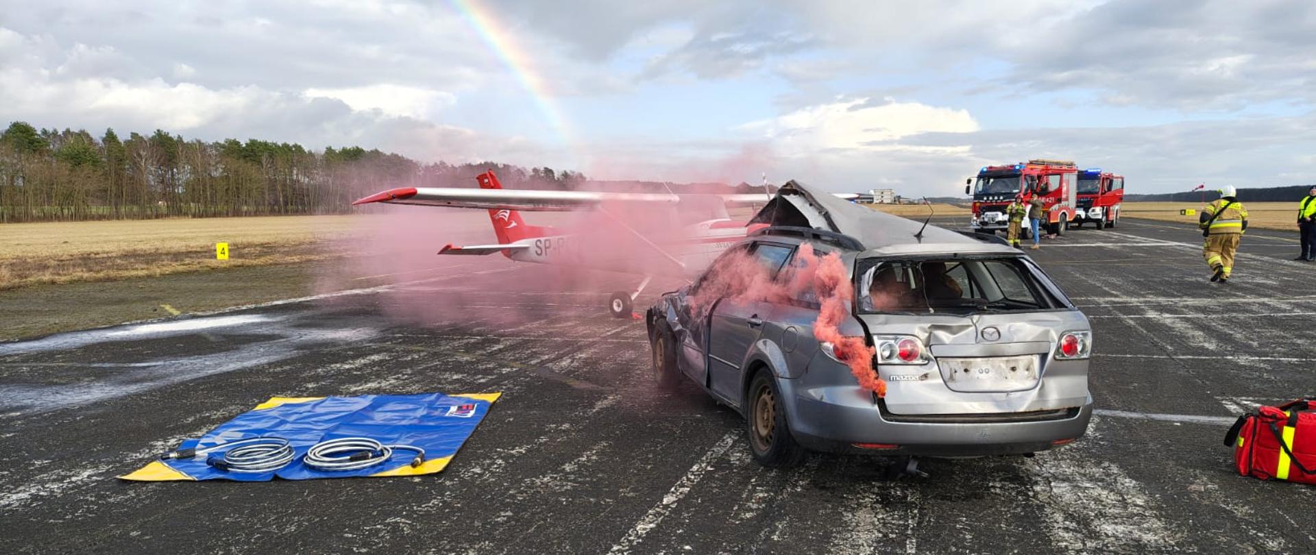 Z samochodu osobowego wydobywa się dym, w tle samolot i pojazdy ratownicze.
