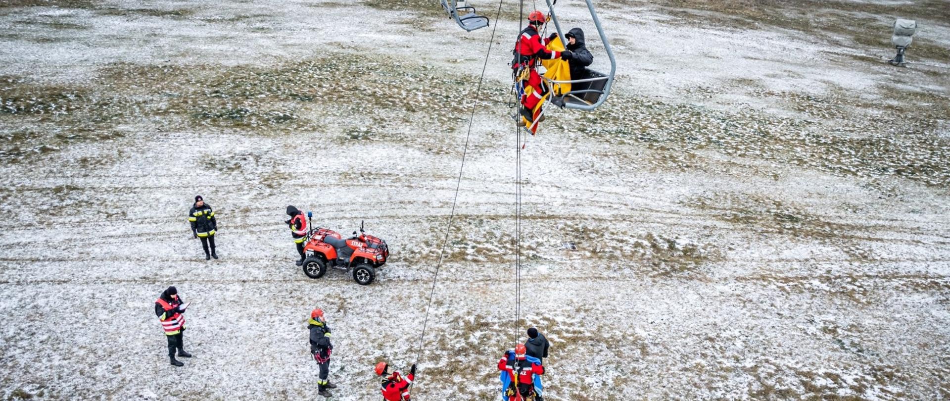 Zdjęcie przedstawia ewakuację osoby poszkodowanej z krzesełkowej kolei linowej, która uległa zatrzymaniu. Na obrazie widocznych jest ośmiu strażaków (w tym osoby funkcyjne podczas ćwiczeń) i pojazd typu Quad na ziemi oraz na wysokości ratownik przystępujący do ewakuacji osoby poszkodowanej z krzesełka kolei przy wykorzystaniu technik linowych