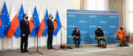 Zdjęcie przedstawia Ministra Spraw Wewnętrznych i Administracji na konferencji prasowej, na której są także przedstawiciele służb - Policji, Straży Granicznej oraz Państwowej Straży Pożarnej z psami.